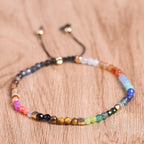 7 chakra bracelet in a close flat lay on warm wood, showing faceted multicolor agate beads, small gold-tone spacers, and the black adjustable cord with two beaded ends for clarity.