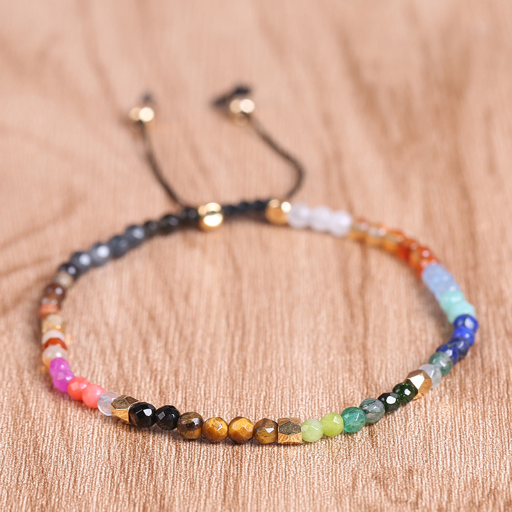 7 chakra bracelet in a close flat lay on warm wood, showing faceted multicolor agate beads, small gold-tone spacers, and the black adjustable cord with two beaded ends for clarity.
