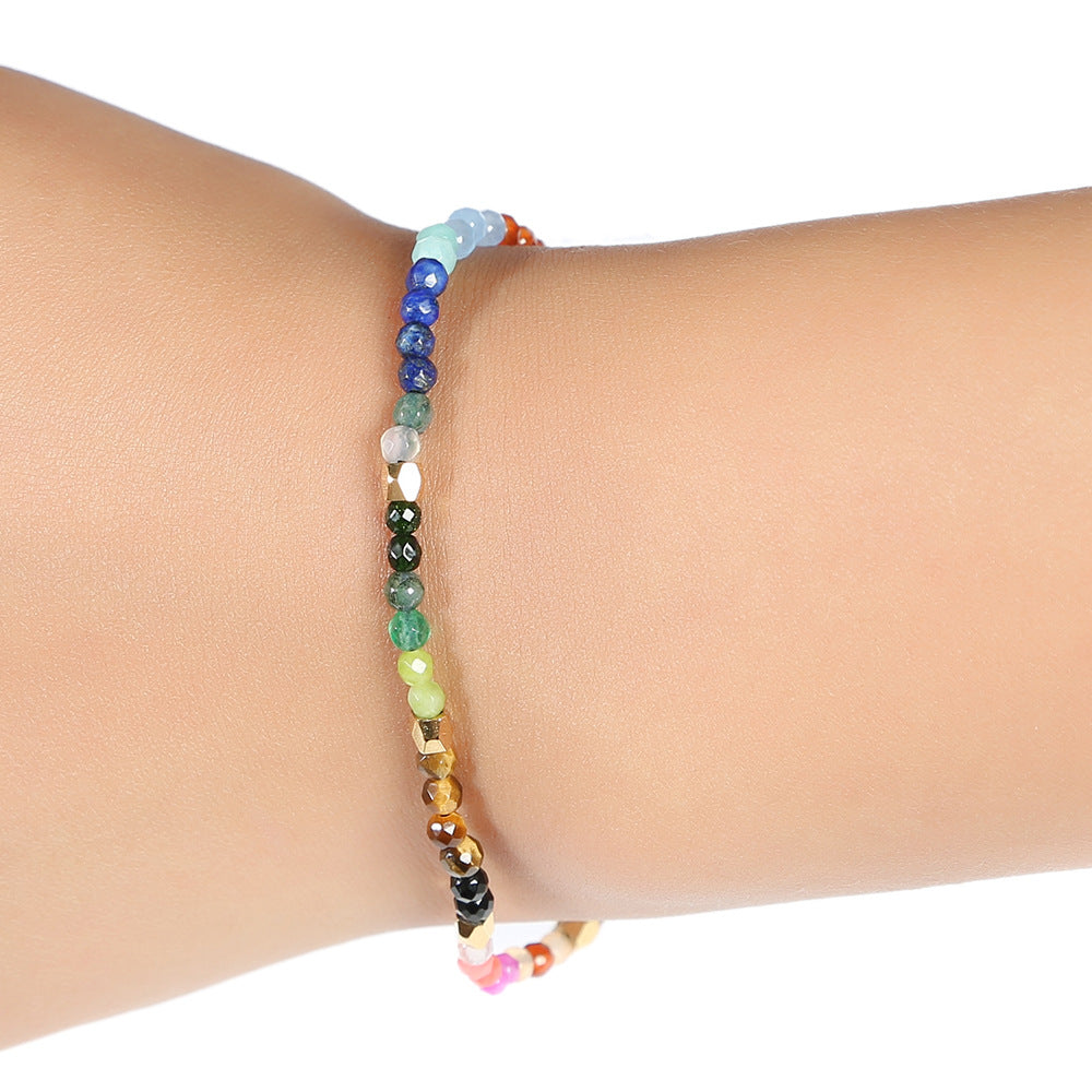 7 chakra bracelet worn on a wrist, showing its slim line of faceted multicolor agate beads, small gold-tone spacers, and black adjustable sliding cord against the white background.