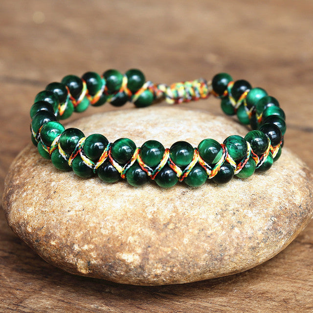 Green adjustable tiger eye bracelet with glossy round beads, multicolor handwoven cord and sliding closure, arranged on a warm natural stone surface to show its woven construction.