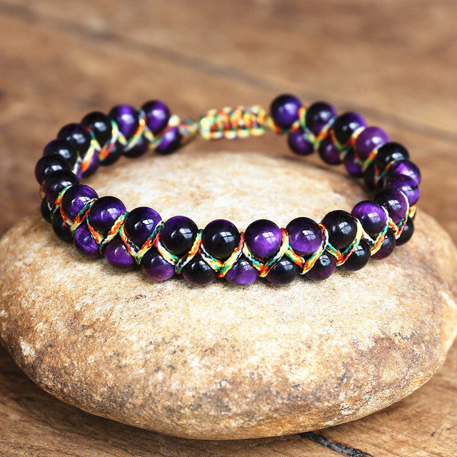 Purple adjustable tiger eye bracelet with shiny round beads, multicolor handwoven cord and sliding closure, arranged on a warm stone surface to clearly show its woven construction.