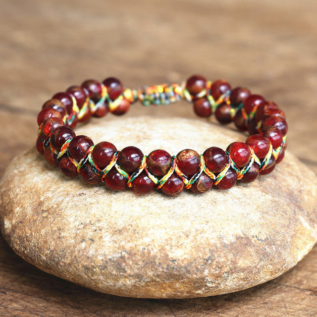 Red adjustable tiger eye bracelet with glossy round beads, multicolor handwoven cord and sliding closure, arranged on a textured stone surface to show its woven construction fully.
