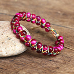 Rose red adjustable tiger eye bracelet with glossy round beads, multicolor handwoven cord and sliding closure, arranged on stones to show the colorful woven construction in detail.