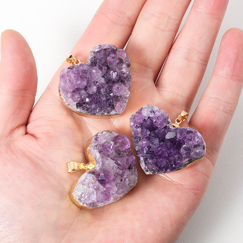 Amethyst heart pendant shown in hand for scale with purple crystal cluster points, irregular natural-style texture, and gold- or silver-color edging with a small jewelry bail loop.