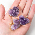 Amethyst heart pendant shown in hand for scale with purple crystal cluster points, irregular natural-style texture, and gold- or silver-color edging with a small jewelry bail loop.