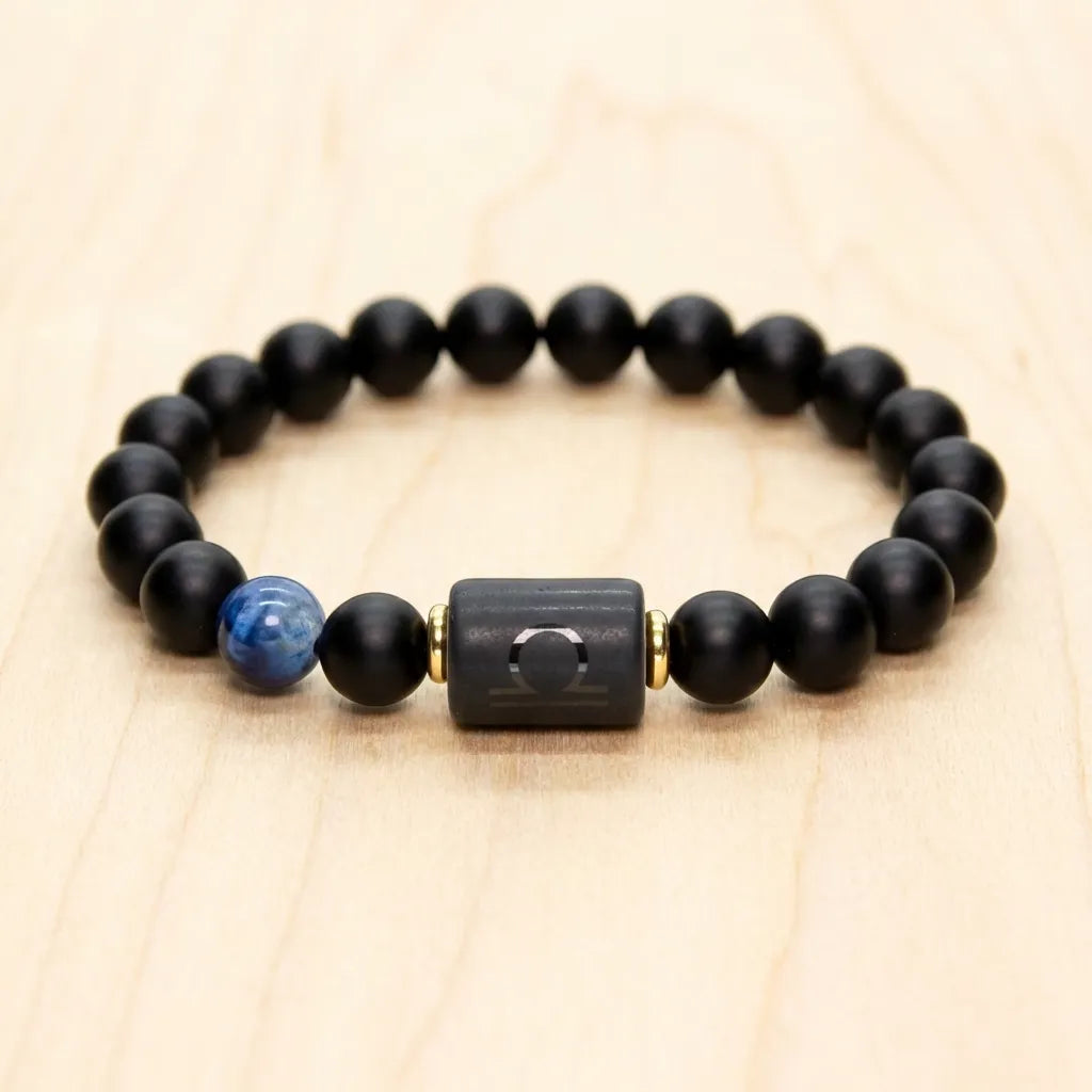 Libra zodiac bracelet with matte black beads, blue accent stone, and engraved center bead on a light wooden surface.