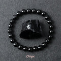 Black onyx bracelet with glossy round beads circling a rough black onyx stone on dark gray fabric, offered in 6 mm and 8 mm sizes plus three wrist lengths on a smooth stretch cord.
