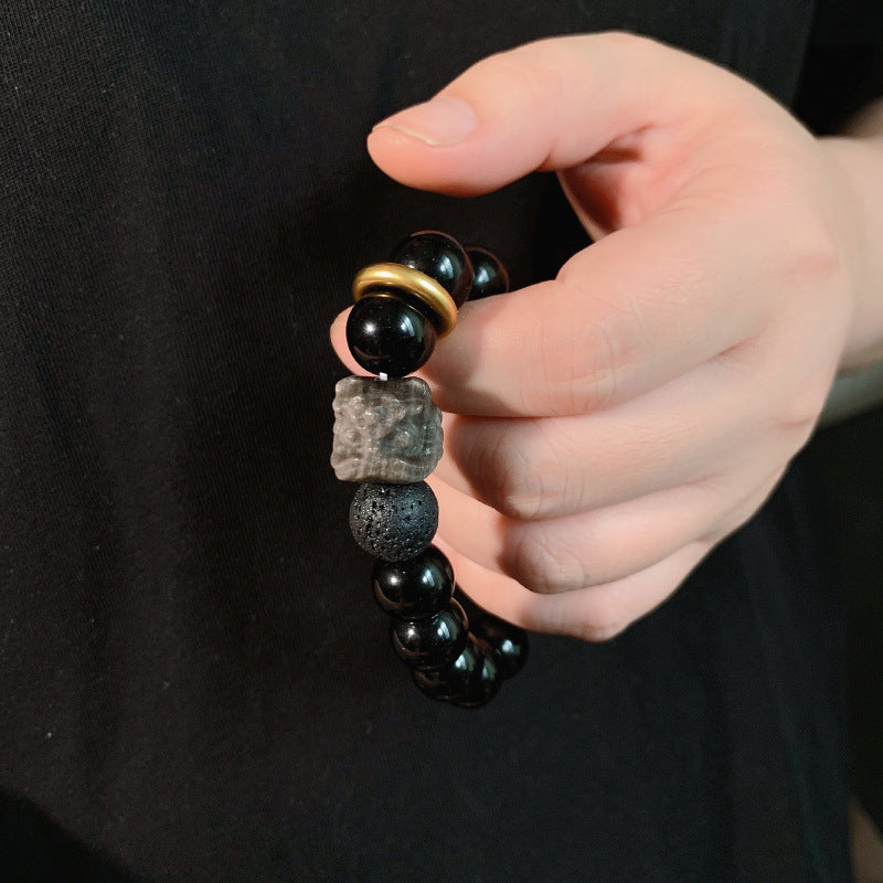 Close wrist view of black stone bracelet with glossy 12mm round beads, smoky gray reflections, one porous accent bead, carved lion-style focal, and gold-colored ring detail in use.
