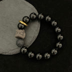 Black stone bracelet flat lay on a dark surface, showing glossy 12mm round beads, smoky gray patterns, a porous accent bead, carved lion-style focal, and gold ring viewed from top.