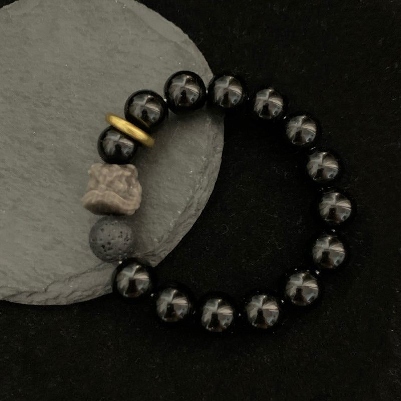 Black stone bracelet flat lay on a dark surface, showing glossy 12mm round beads, smoky gray patterns, a porous accent bead, carved lion-style focal, and gold ring viewed from top.