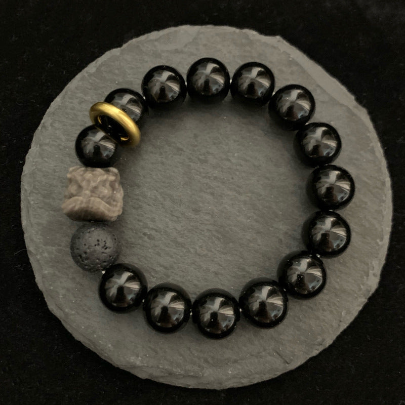 Black stone bracelet displayed on gray stones, with glossy 12mm round beads, smoky gray patterns, one porous accent bead, carved lion-style focal, and gold-colored ring from above.