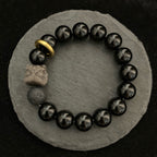 Black stone bracelet displayed on gray stones, with glossy 12mm round beads, smoky gray patterns, one porous accent bead, carved lion-style focal, and gold-colored ring from above.