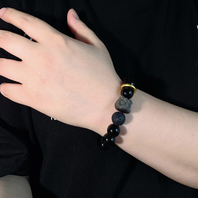 Black stone bracelet worn on the wrist, showing glossy 12mm round beads, a carved lion-style focal bead, one porous accent bead, and a thick gold-colored ring detail in clear view.
