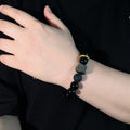 Black stone bracelet worn on the wrist, showing glossy 12mm round beads, a carved lion-style focal bead, one porous accent bead, and a thick gold-colored ring detail in clear view.