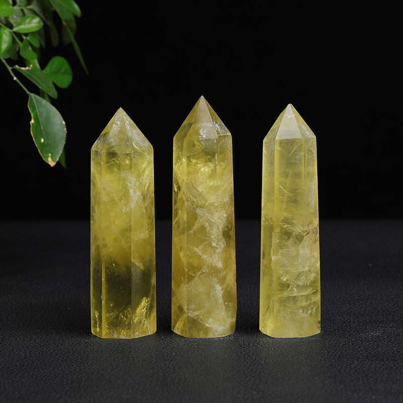 Citrine crystal tower trio with polished yellow faces, pointed tops, and clear inner lines arranged upright on a black surface beneath a small green plant at upper left, in studio.