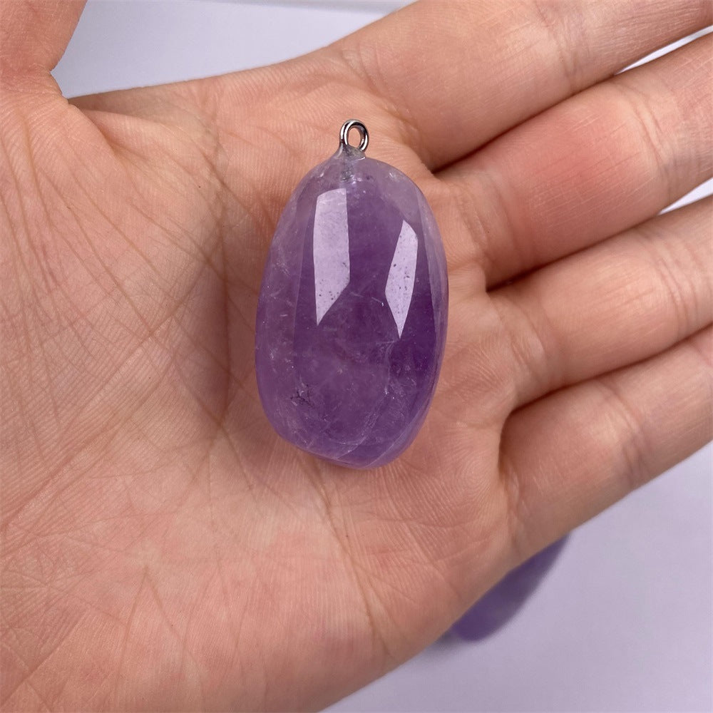 Freeform amethyst pendant shown in hand for scale with a polished purple pebble shape, visible tonal bands and inclusions, plus one small silver-color hanging loop at the top edge.
