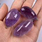 Freeform amethyst pendant trio held in one hand with polished purple pebble shapes, varied violet color, visible internal inclusions, and tiny silver-color hanging loops for scale.
