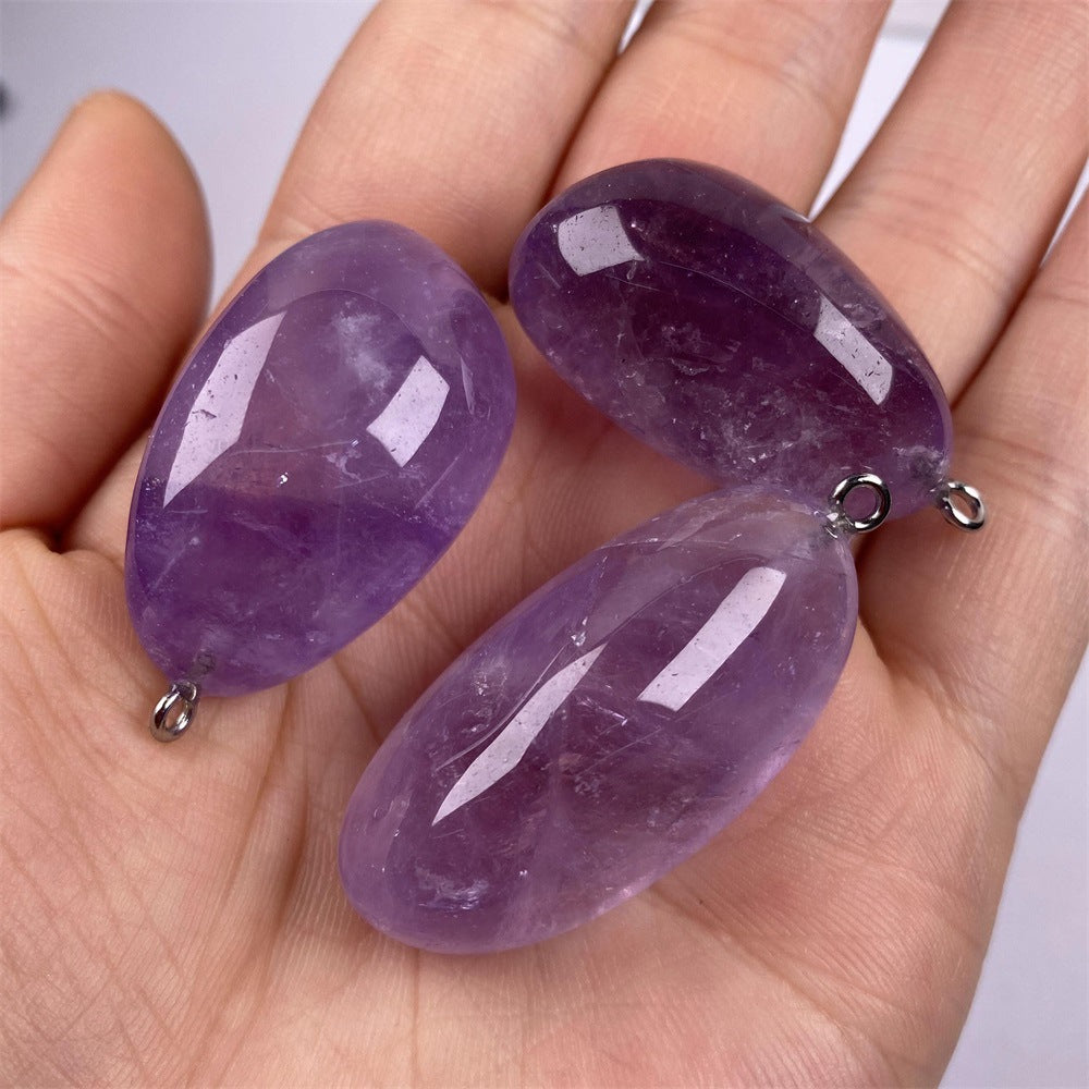 Freeform amethyst pendant trio held in one hand with polished purple pebble shapes, varied violet color, visible internal inclusions, and tiny silver-color hanging loops for scale.