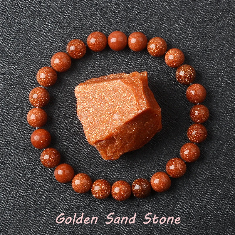 Goldstone bracelet with polished reddish-brown beads and copper-gold sparkle, arranged with a raw goldstone piece on dark gray textured fabric, offered in 6 mm and 8 mm bead sizes.