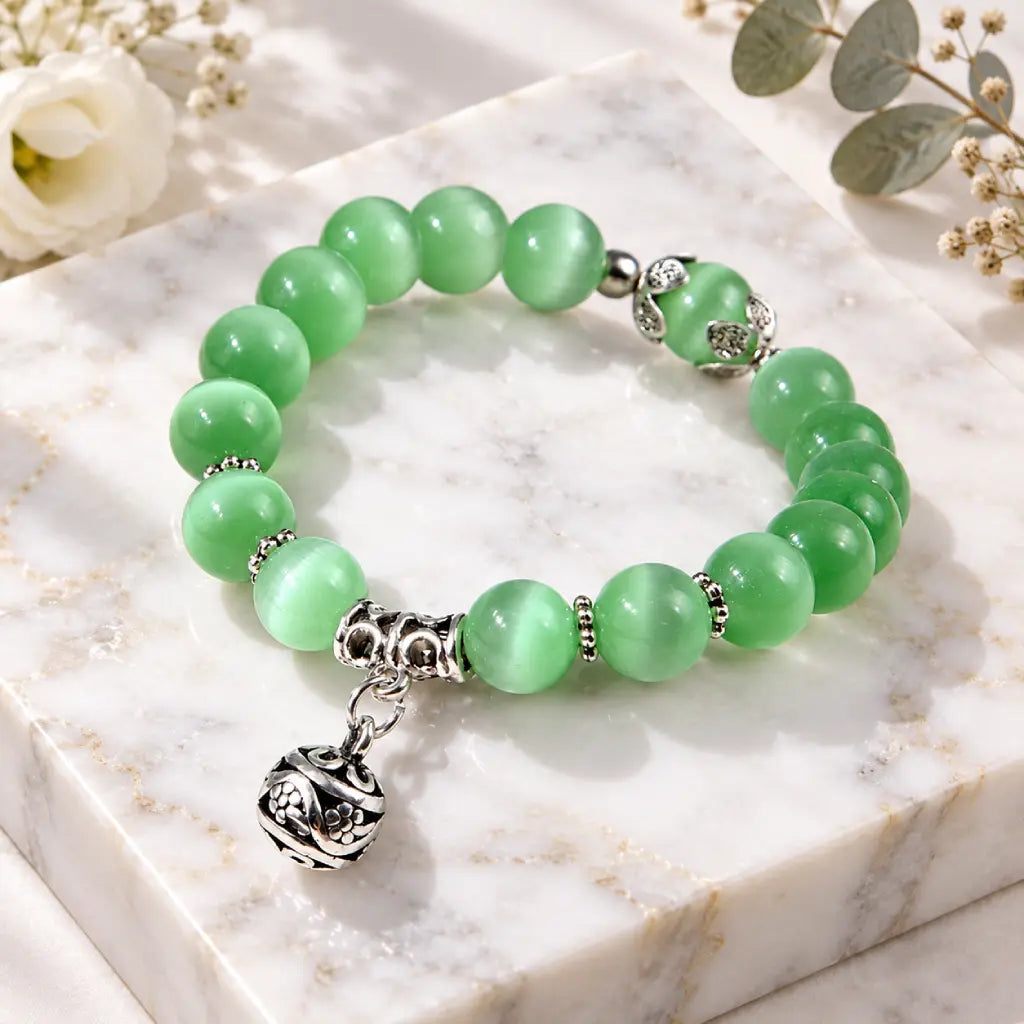 Green catseye bracelet featuring polished green beads and dangling silver floral charm, styled on marble for effortless elegance.
