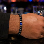 Close-up of a wrist wearing a black beaded bracelet with a blue bead, against a blurred bar background.