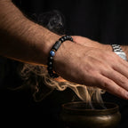Libra zodiac bracelet worn on a wrist above a smoking bowl, featuring black beads and a blue accent stone.