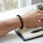Person writing in a notebook with a pen, wearing a black beaded bracelet on a light surface.