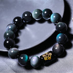 Rainbow obsidian bracelet in a detailed circular flat lay, showing polished black beads with green, blue, and purple sheen, plus a black barrel accent with decorative gold symbols.