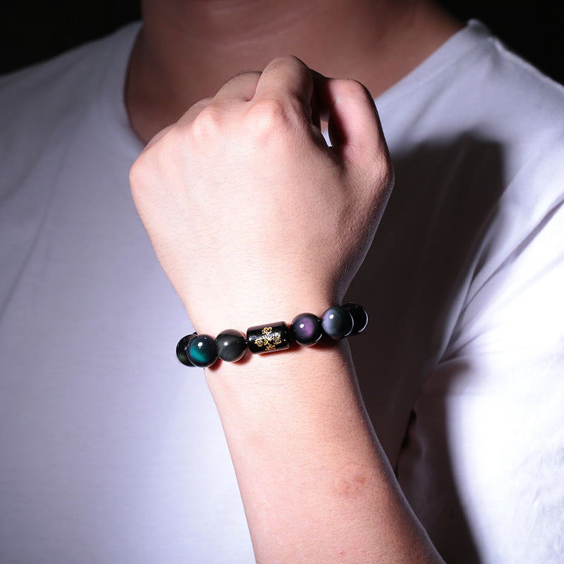 Rainbow obsidian bracelet worn on a wrist in a close lifestyle view, showing glossy dark round beads, subtle multicolor sheen, and a black barrel accent with detailed gold symbols.