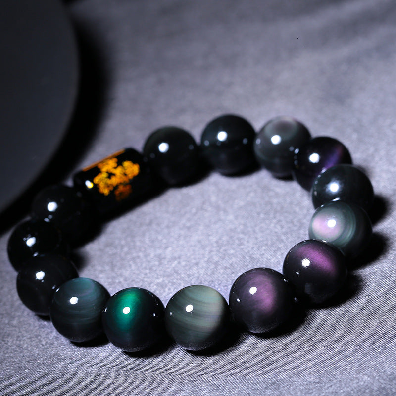 Rainbow obsidian bracelet arranged in a circle on charcoal fabric with polished black beads reflecting green, blue, and purple sheen around a black barrel accent with gold symbols.
