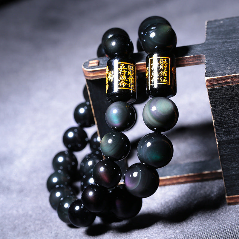 Rainbow obsidian bracelet pair with glossy black beads showing green and purple sheen, plus black barrel accents with gold symbols, arranged on a black display for size comparison.