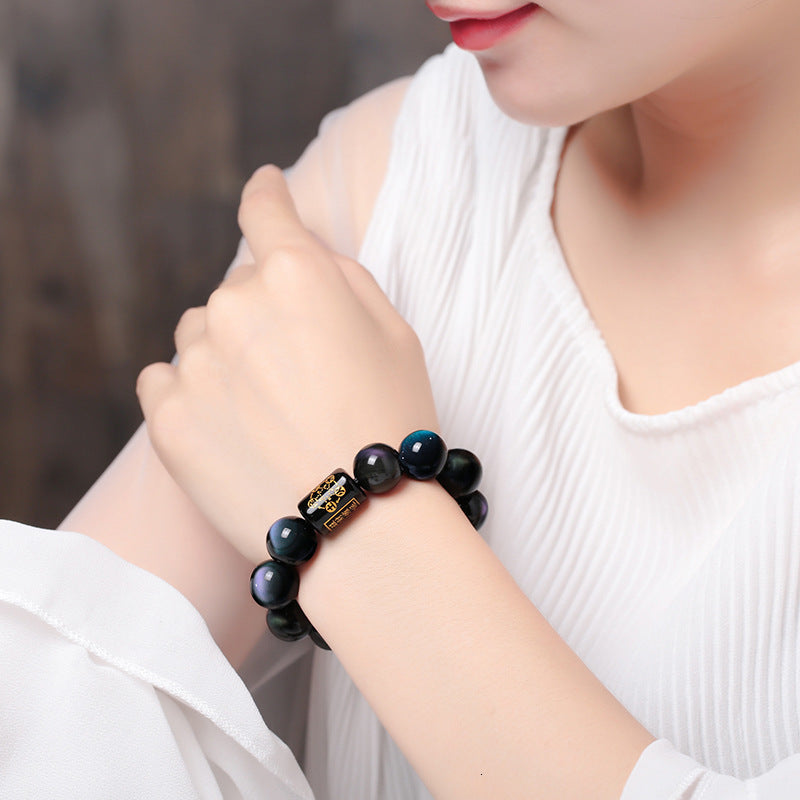 Rainbow obsidian bracelet worn on a woman's wrist showing polished dark beads with subtle green and blue sheen and a black barrel accent with gold symbols against a neutral outfit.