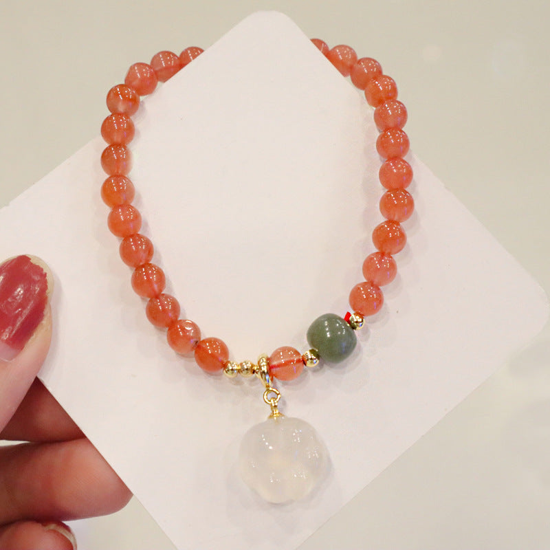 Red agate bracelet with orange-red round beads, a green accent bead, gold-tone spacer details, and a white cat-paw charm, shown on a white jewelry card held in a hand in full view.