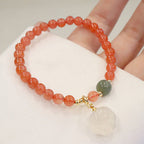 Red agate bracelet with translucent orange-red round beads, a green accent bead, gold-tone spacers, and a white cat-paw charm, displayed over soft white fabric in a hand-held view.