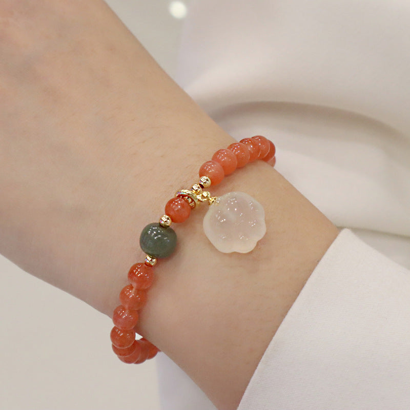 Red agate bracelet worn around the wrist, showing translucent orange-red round beads, one green accent bead, gold-tone spacers, and an ivory cat-paw charm in a soft indoor setting.