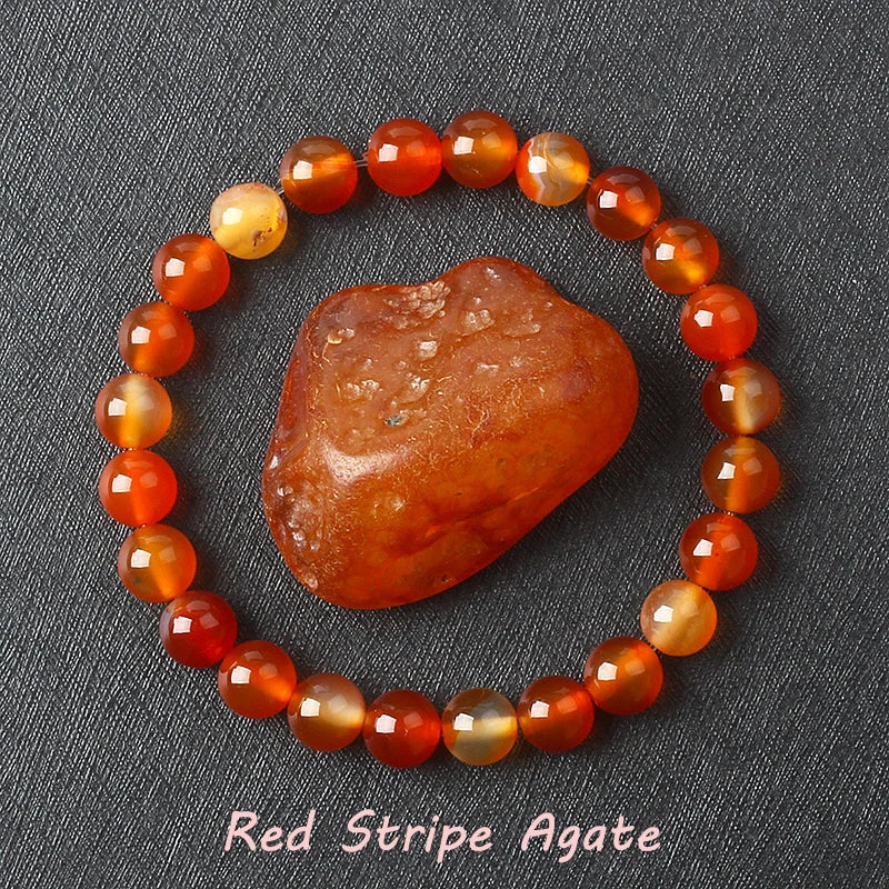Red stripe agate bracelet with polished red, orange, amber, and cream round beads, shown around a matching raw stone on dark gray fabric, available in 6 mm and 8 mm bead sizes too.