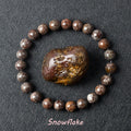 Snowflake Obsidian bracelet with dark brown beads and white crystal patterns surrounding a raw obsidian specimen on dark background