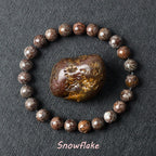Snowflake Obsidian bracelet with dark brown beads and white crystal patterns surrounding a raw obsidian specimen on dark background