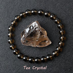 Tea Crystal bracelet with round smoky brown beads beside a raw matching crystal stone on dark fabric