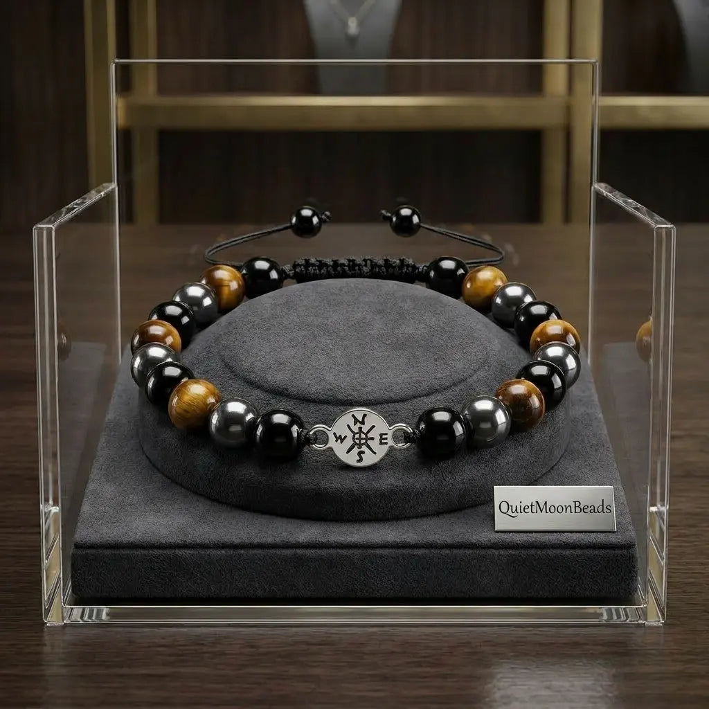 Beaded bracelet with a compass charm on a gray stand inside a clear display case, with 'Quiet Moon Beads' branding.