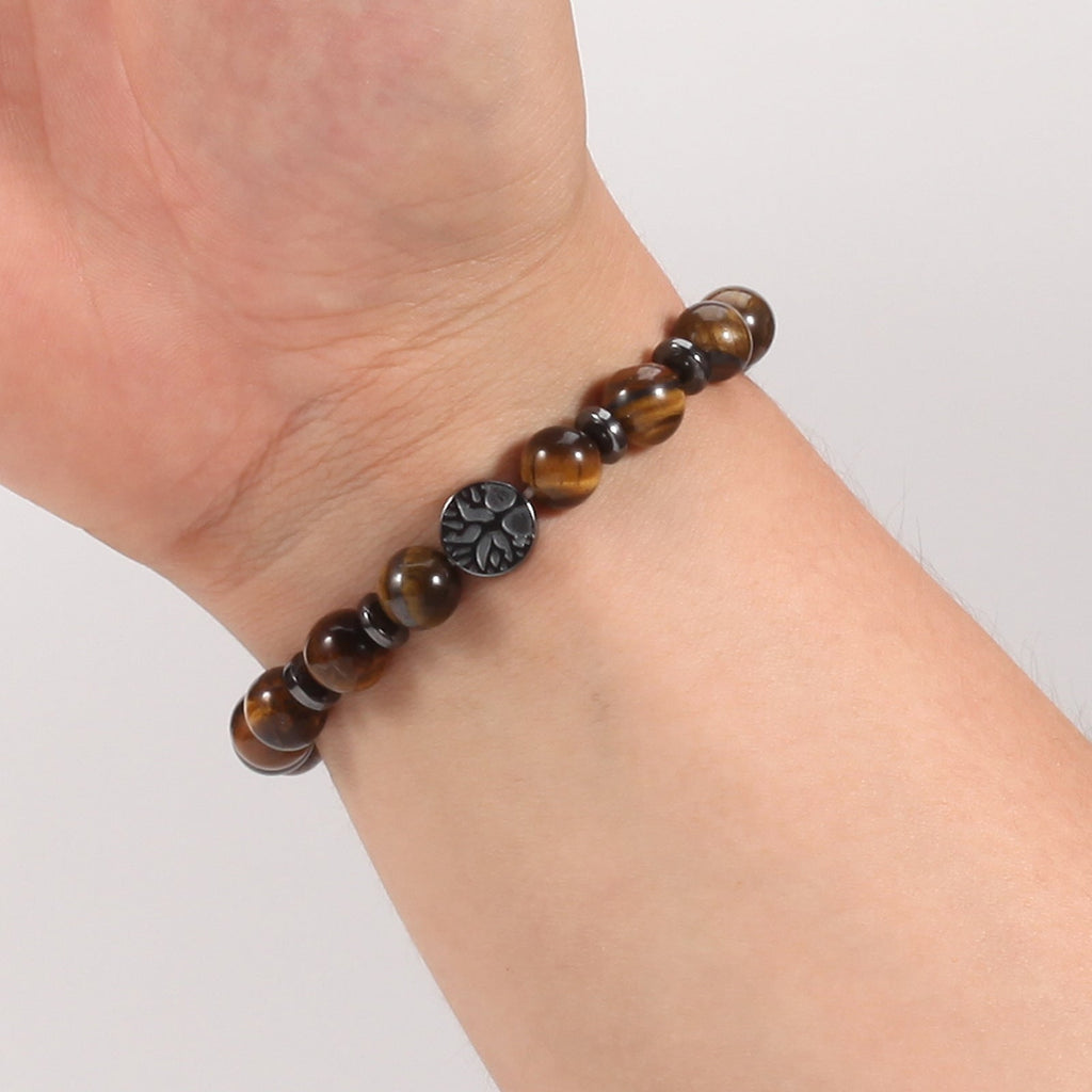 Tree of life stone bracelet worn on a wrist in the Tiger Eye option, with glossy brown 8mm beads, black spacer rings, and a round dark tree-design focal bead on a white background.