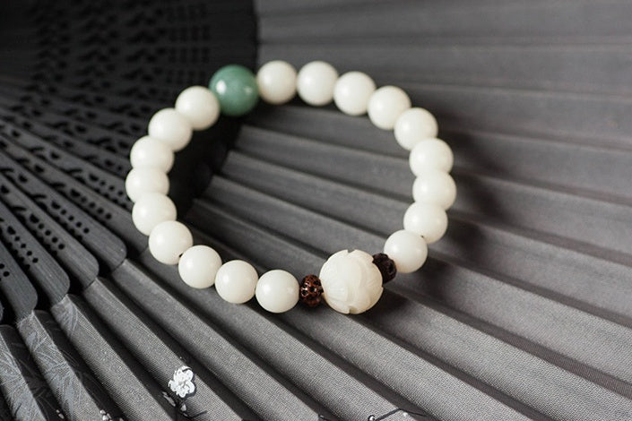 White bodhi seed bracelet arranged on dark bamboo slats with cream round beads, one green accent bead, dark carved spacers, and a larger white lotus style focal bead in fair light.