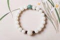 White bodhi seed bracelet displayed flat with smooth round ivory-color beads, one green accent bead, two dark carved spacers, and a larger carved white lotus-style focal bead here.