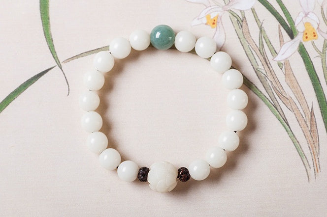 White bodhi seed bracelet displayed flat with smooth round ivory-color beads, one green accent bead, two dark carved spacers, and a larger carved white lotus-style focal bead here.