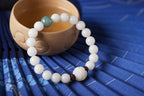 White bodhi seed bracelet beside a wooden tea bowl on blue fabric showing pale round beads, one green accent bead, dark spacers, and a carved white lotus-style focal bead close by.