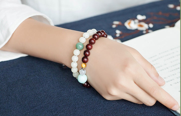 White bodhi seed bracelet worn beside brown and pastel bead strands, showing its smooth pale round beads and carved white lotus-style focal bead near an open book in a quiet scene.