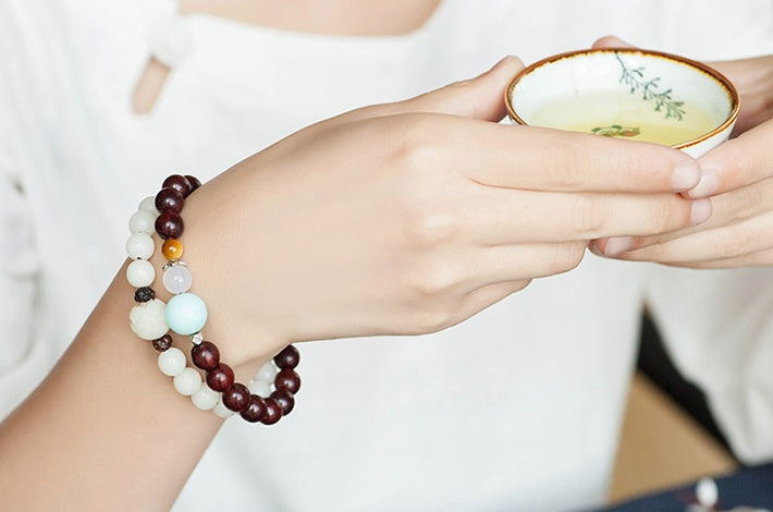 White bodhi seed bracelet worn in a layered wrist stack, showing cream round beads and a carved white focal bead beside brown and pastel bead bracelets during tea under soft light.